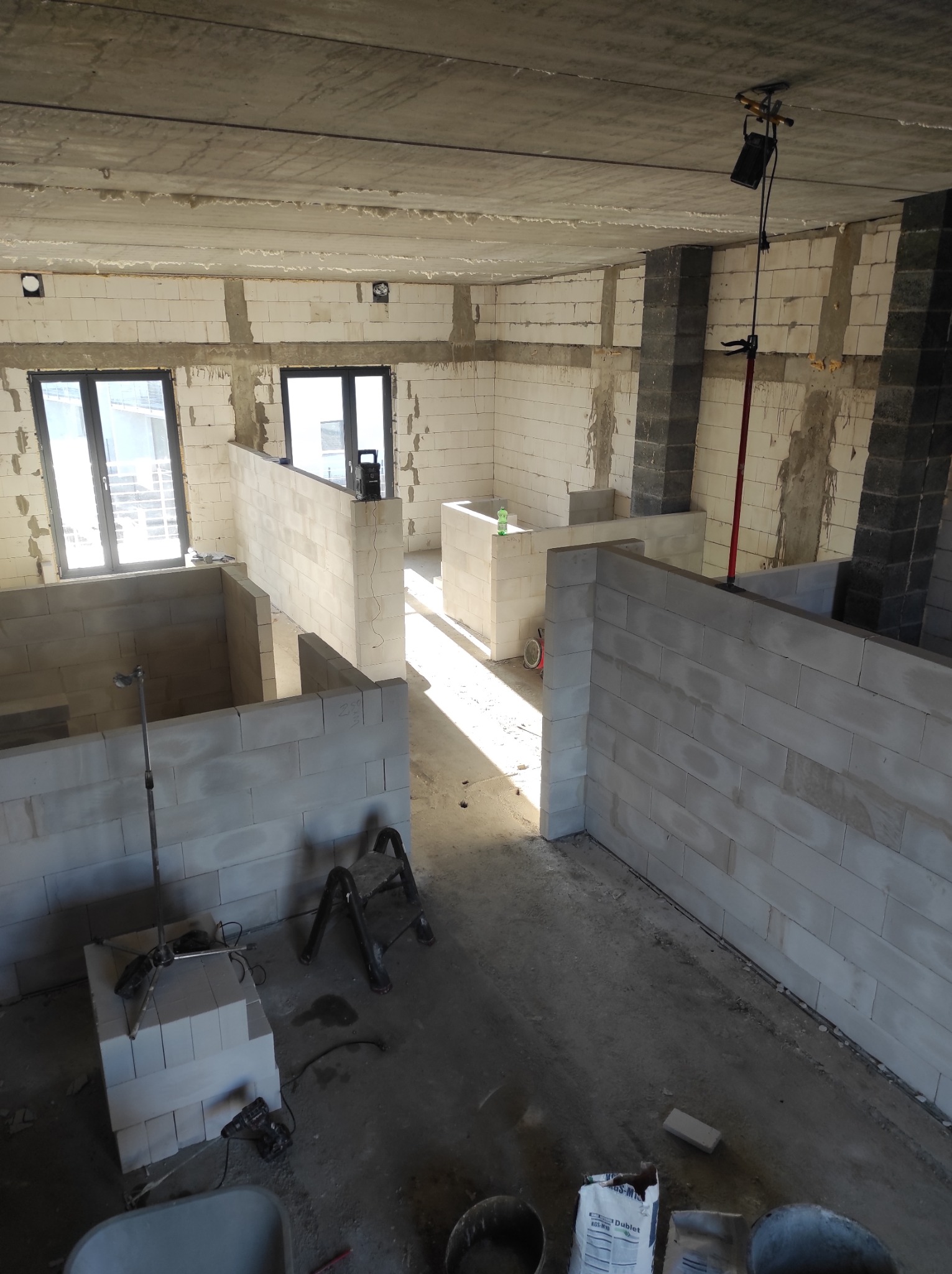Wnętrze w trakcie budowy ścian działowych z betonu komórkowego, widoczne narzędzia i materiały budowlane, surowy betonowy strop, okna w tle.