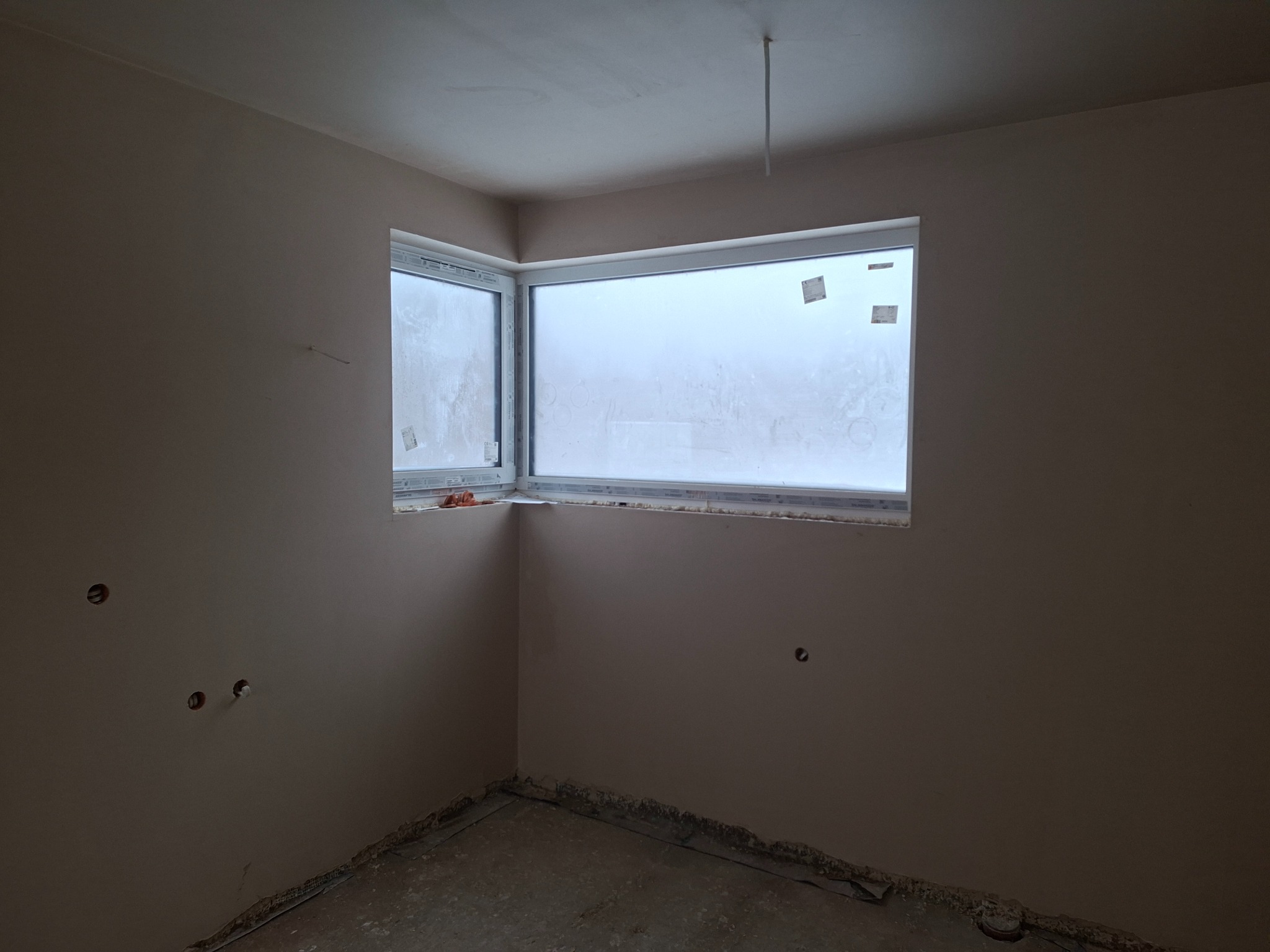 Pokój po nałożeniu gładzi gipsowej. Widoczne gładkie ściany w jasnym odcieniu, okno z folią ochronną i wystające kable elektryczne. Podłoga przygotowana do dalszych prac.