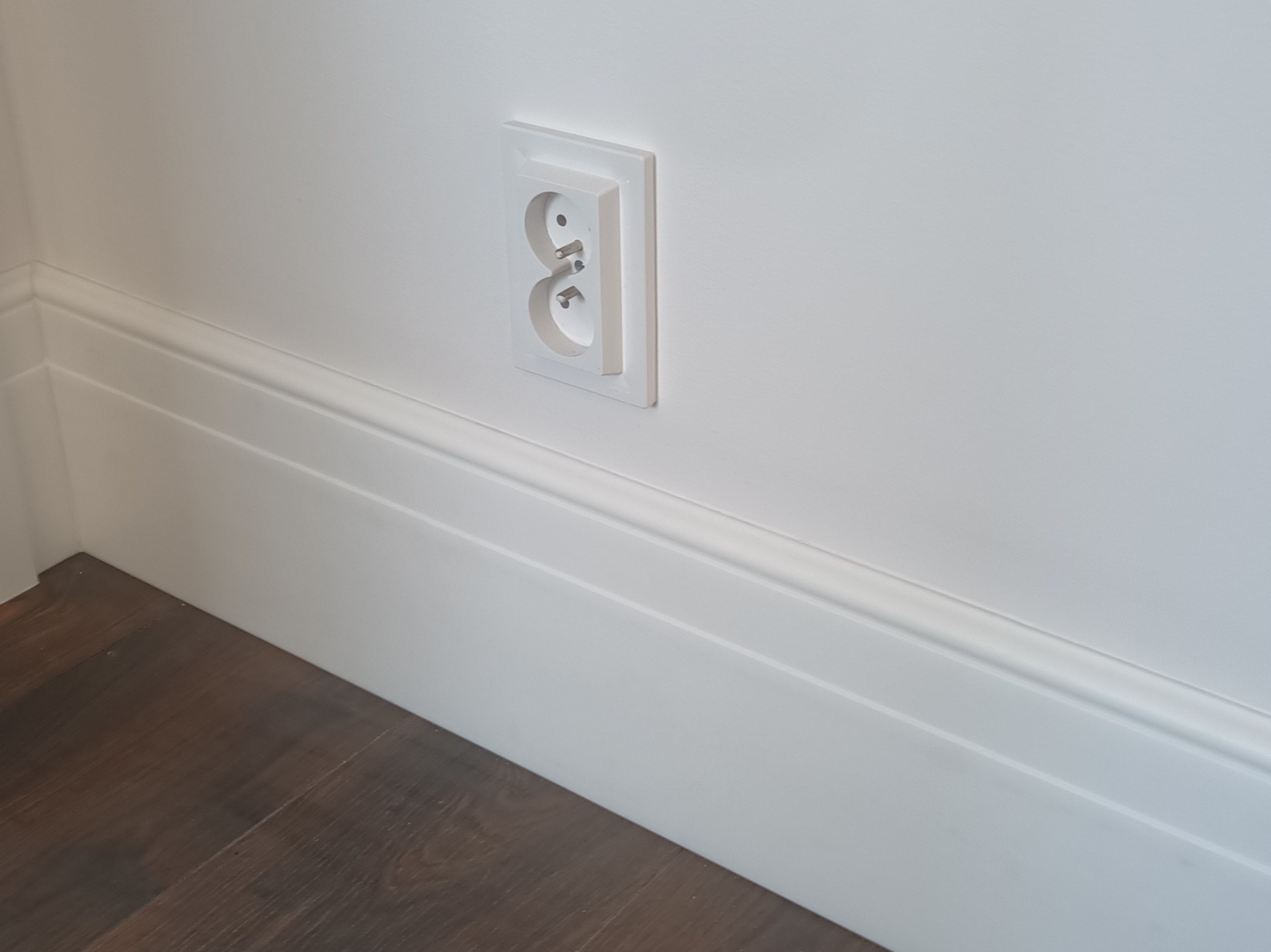 Białe gniazdko elektryczne z bolcem, zamontowane w ścianie nad białą listwą przypodłogową i ciemną podłogą. Minimalistyczny design wnętrza.