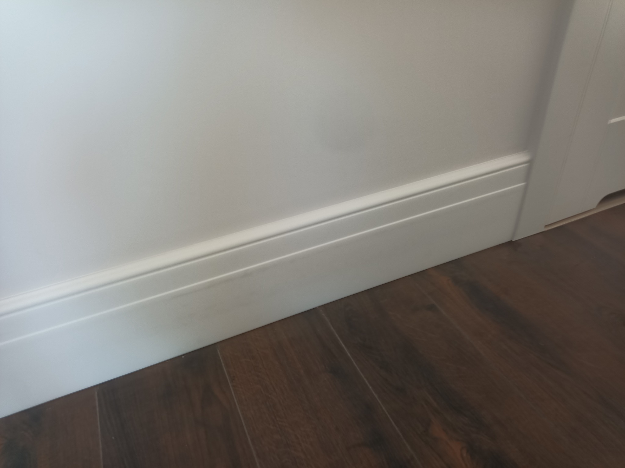 Biała listwa przypodłogowa z delikatnym profilem, zamontowana przy ścianie pomalowanej na biało, z widoczną podłogą z paneli imitujących ciemne drewno.