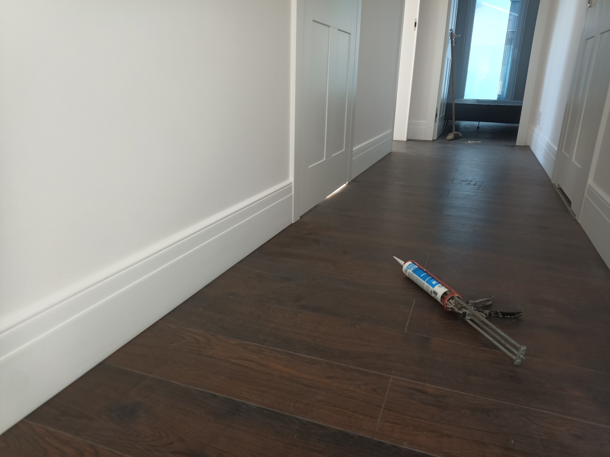 Białe listwy przypodłogowe zamontowane wzdłuż ściany na ciemnej podłodze. Pistolet do silikonu leży na podłodze. Widok na korytarz z drzwiami.