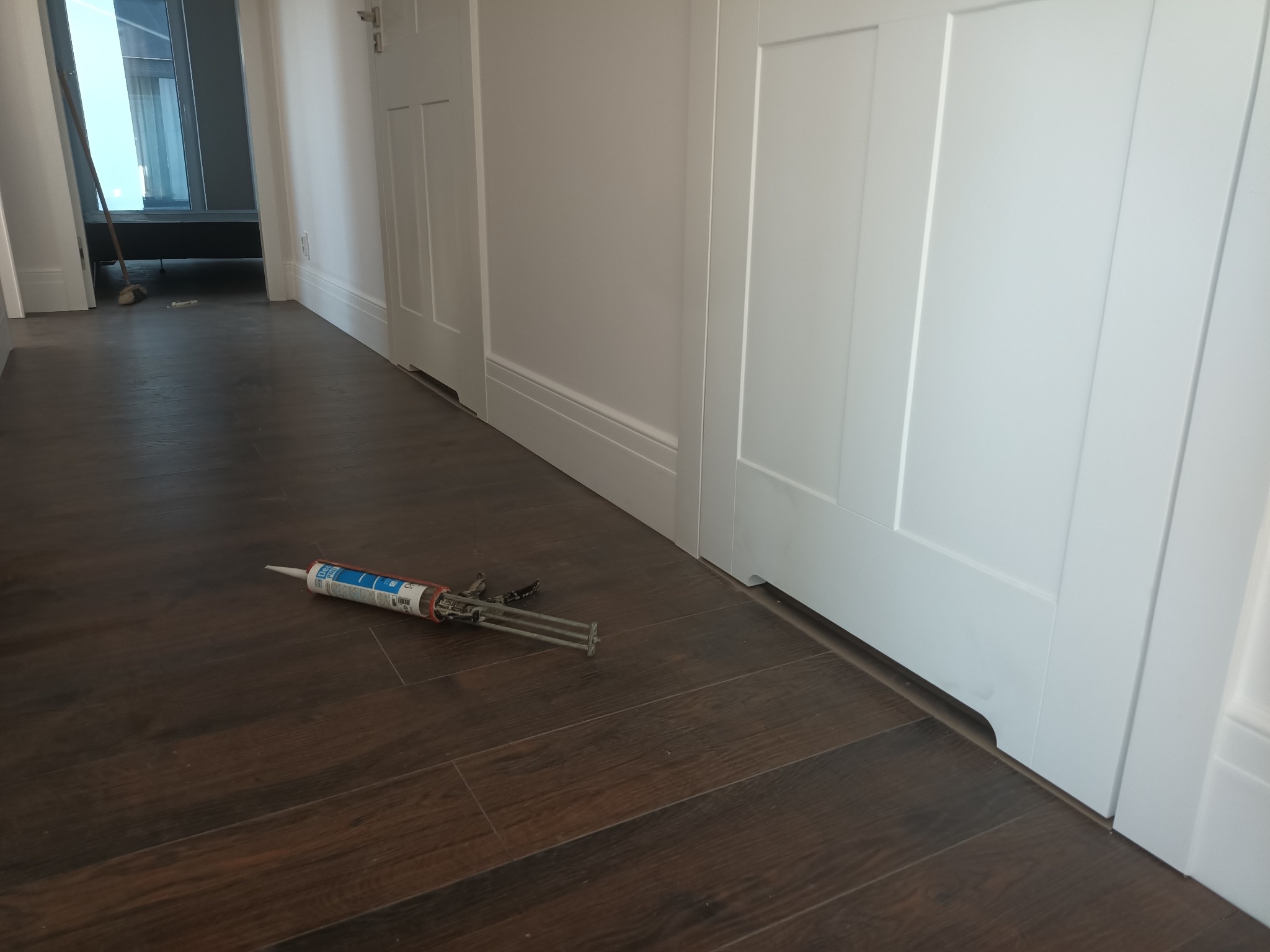 Białe, panelowe drzwi wewnętrzne po montażu, widoczne z poziomu podłogi z ciemnych paneli. Na podłodze leży pistolet do silikonu.