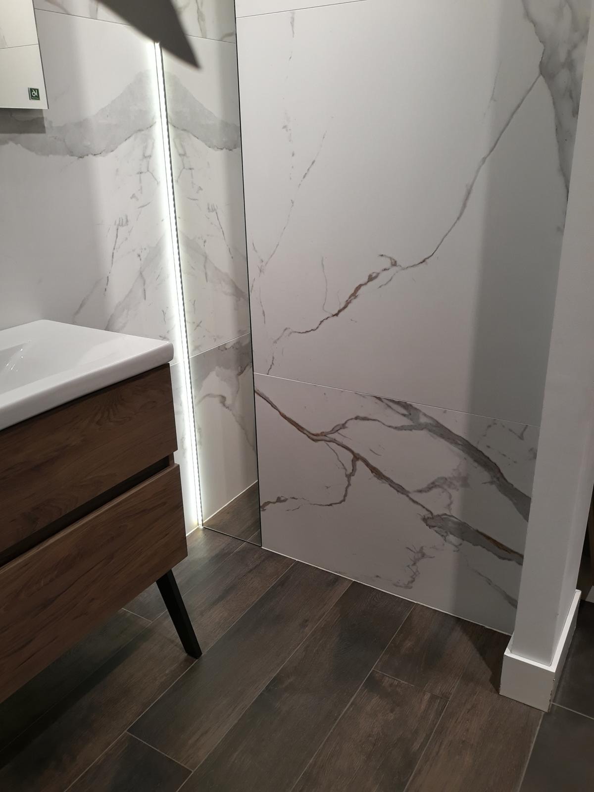 Nowoczesna łazienka z drewnianą szafką pod umywalką, płytkami imitującymi marmur na ścianach i podświetleniem LED w lustrze. Ciemna podłoga z paneli.