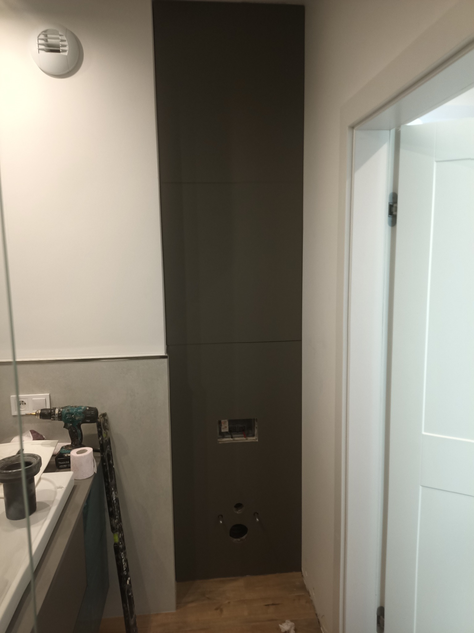 Montaż paneli ściennych w łazience, widoczne narzędzia i otwory na instalacje. Szare panele, białe ściany i drzwi. Prace wykończeniowe w toku.