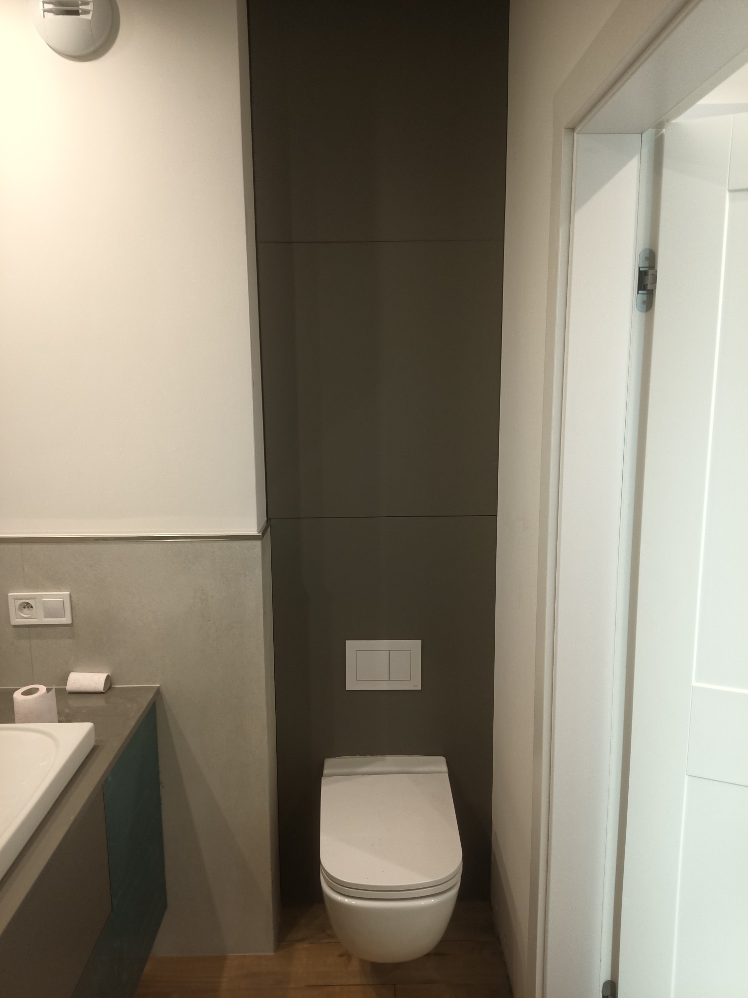 Nowoczesna toaleta w łazience z szarą zabudową, białym sedesem i ukrytą spłuczką. Widoczna biała ściana i fragment umywalki. Minimalistyczny design.