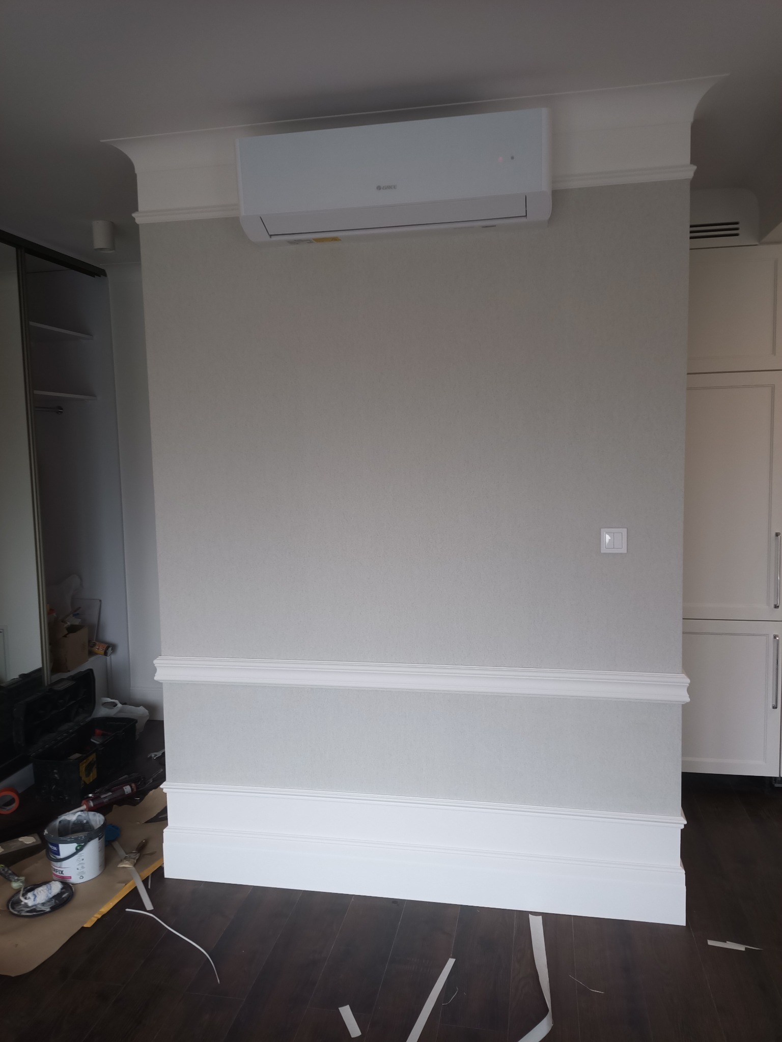 Ściana po remoncie z klimatyzatorem, ozdobnymi listwami przypodłogowymi i sufitowymi. Widoczne narzędzia malarskie i zabezpieczenia podłogi. Parkiet w ciemnym odcieniu.