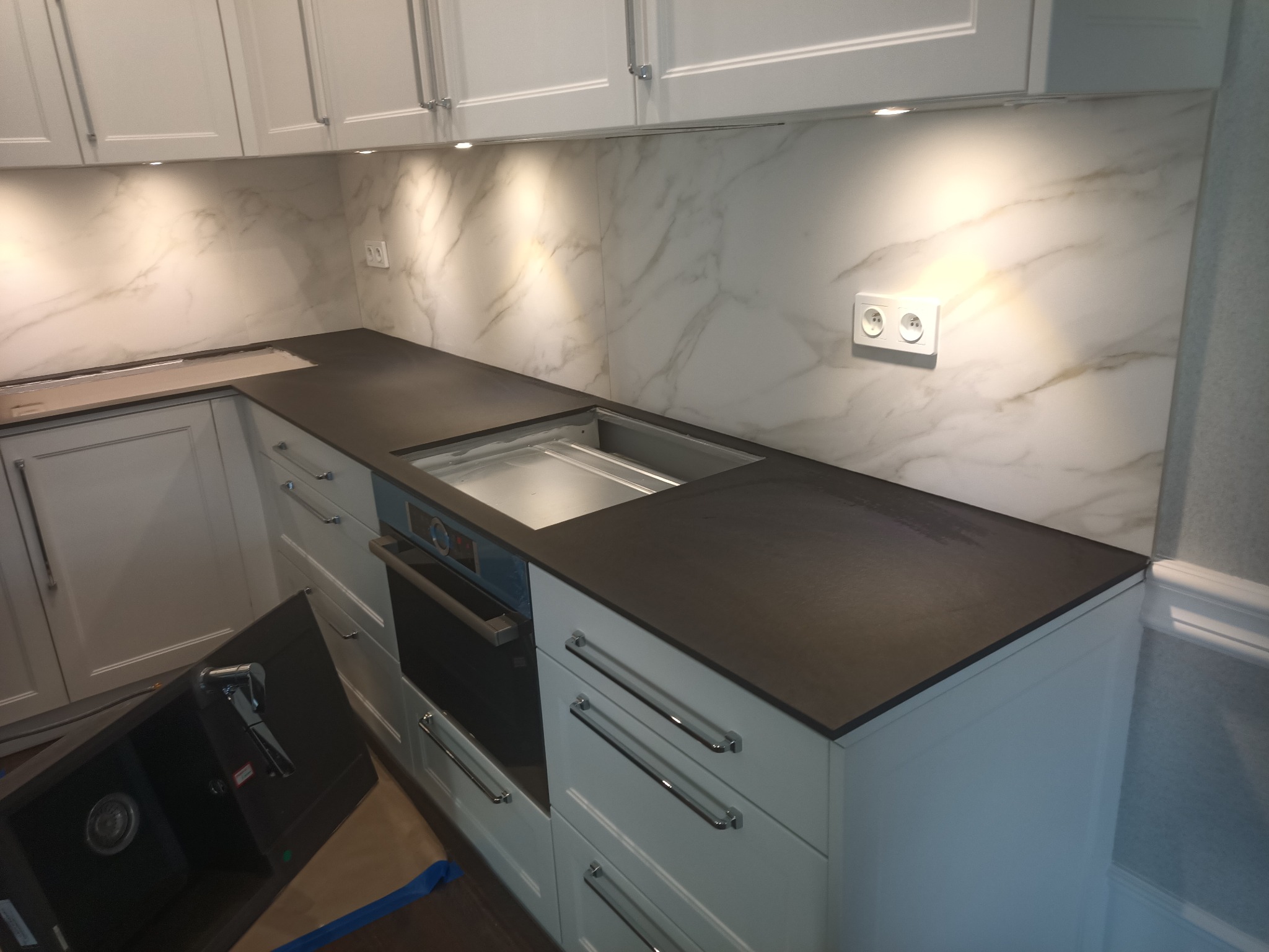 Nowoczesna kuchnia z białymi szafkami, ciemnym blatem i marmurową ścianą. W tle widoczne oświetlenie podszafkowe i gniazdko elektryczne. Częściowo zamontowany zlew.
