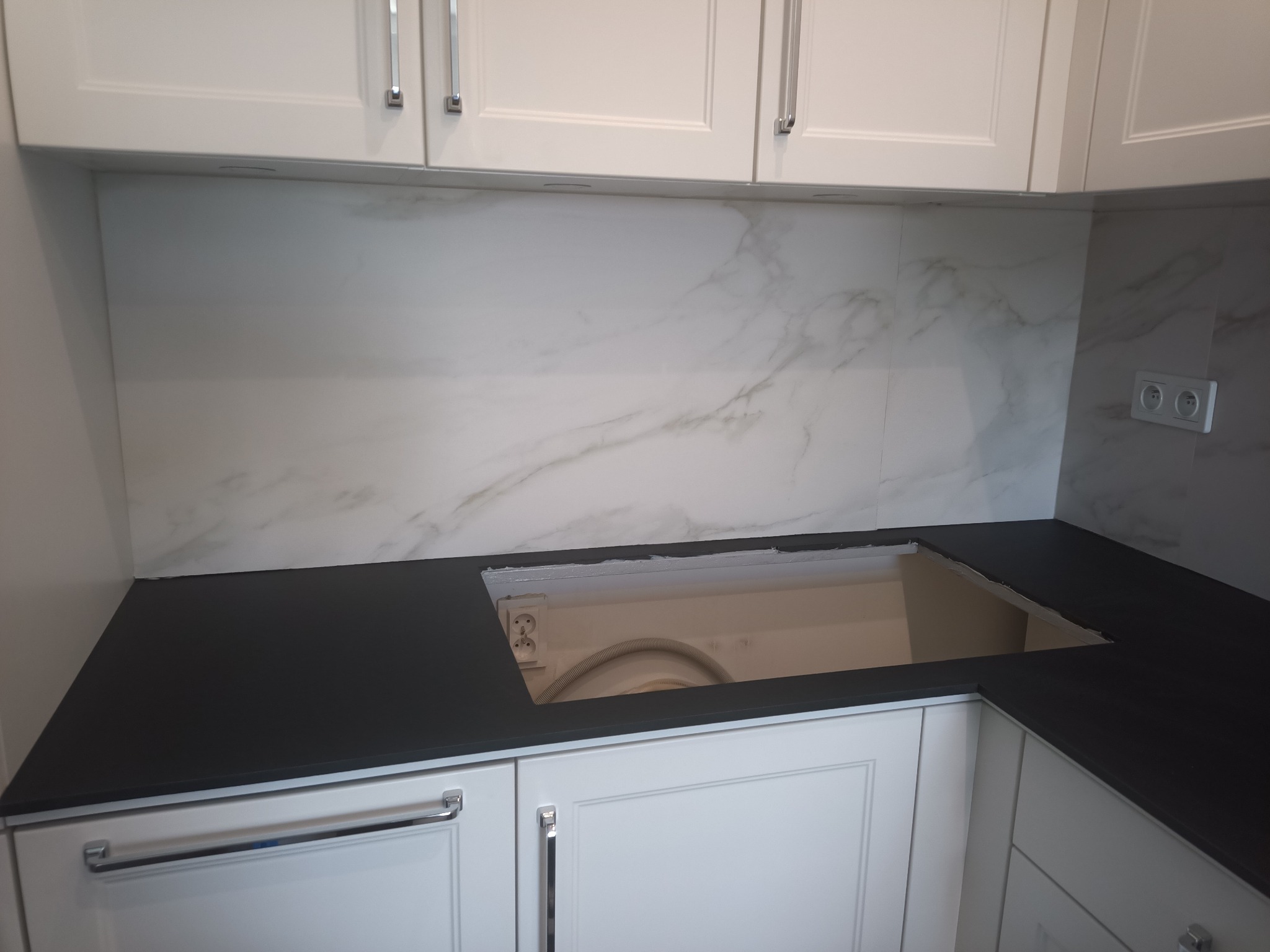Biała zabudowa kuchenna z czarnym blatem i marmurowym panelem ściennym. Widoczny otwór w blacie pod zlew, gniazdko elektryczne. Ujęcie z bliska, detale wykończenia.