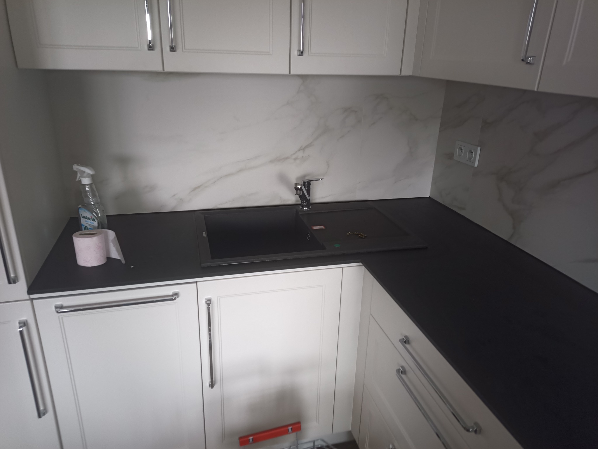 Białe szafki kuchenne z ciemnym blatem i marmurowym panelem ściennym. Widoczny zlew, bateria i akcesoria. Ujęcie z perspektywy narożnika kuchni.