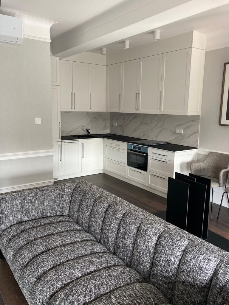 Nowoczesna, jasna kuchnia z białymi szafkami i marmurowym backsplashiem, widziana z perspektywy salonu z szarą sofą. Minimalistyczny design, ciemna podłoga.