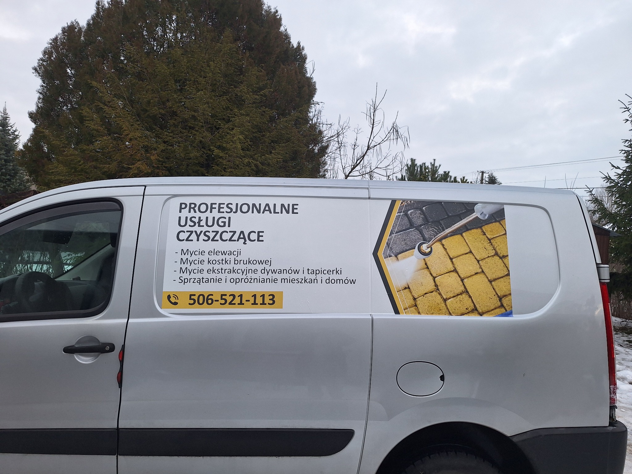 Srebrny samochód dostawczy z reklamą usług czyszczących: mycie elewacji, kostki brukowej, dywanów i tapicerki. Widoczny numer telefonu. Tło: drzewa i pochmurne niebo.