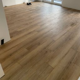 Perfect Wnętrza - Ukończona podłoga z paneli imitujących drewno w jasnym, przestronnym pokoju z białymi ścianami i dużym oknem balkonowym. Widoczne detale wykończenia.