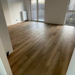 Perfect Wnętrza - Jasne wnętrze z drewnopodobną podłogą, grzejnikiem i balkonem z widokiem na drzewa. Świeżo wykończone, minimalistyczne pomieszczenie z naturalnym światłem.