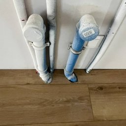 Perfect Wnętrza - Pionowe rury, częściowo pomalowane na biało i niebiesko, przymocowane do ściany nad drewnianą podłogą. Widoczne ślady farby na podłodze i rurach.