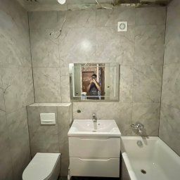 Perfect Wnętrza - Wyremontowana łazienka z szarymi płytkami imitującymi marmur, białą armaturą i lustrem. Widoczne niedokończone prace na suficie i odbicie osoby robiącej zdjęcie.