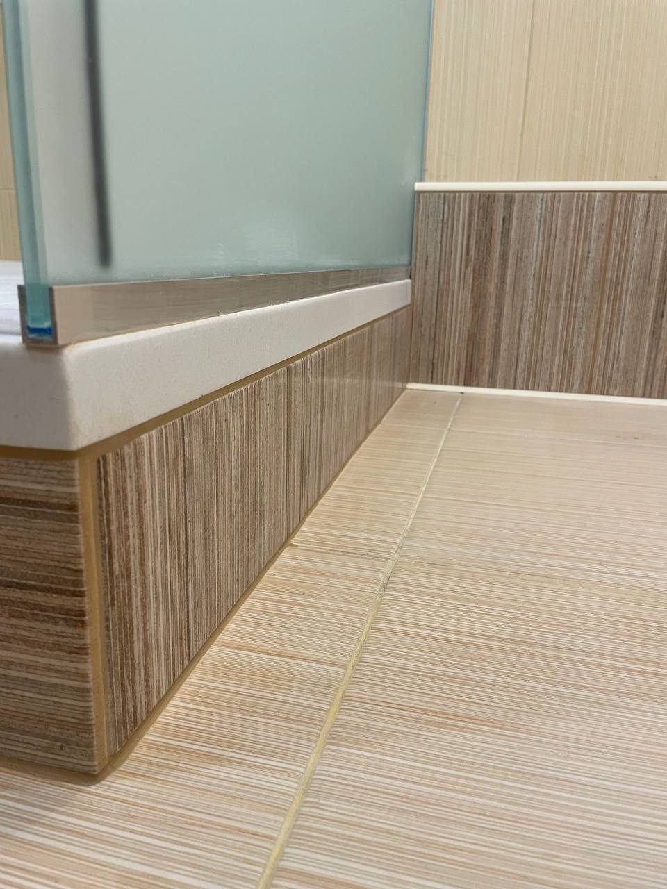 Detal wykończenia łazienki: beżowe płytki podłogowe i ścienne z delikatnym wzorem drewna, biały cokół i szklana ścianka kabiny prysznicowej z metalowym profilem.