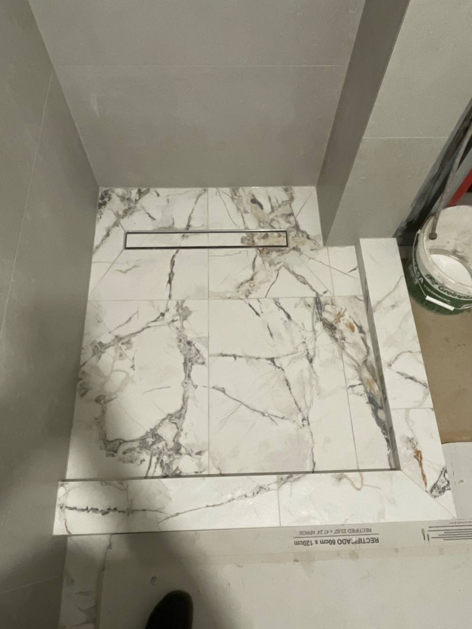 Ułożone płytki imitujące marmur z odpływem liniowym w kabinie prysznicowej. Widoczny fragment opakowania płytek i wiaderko z zaprawą.