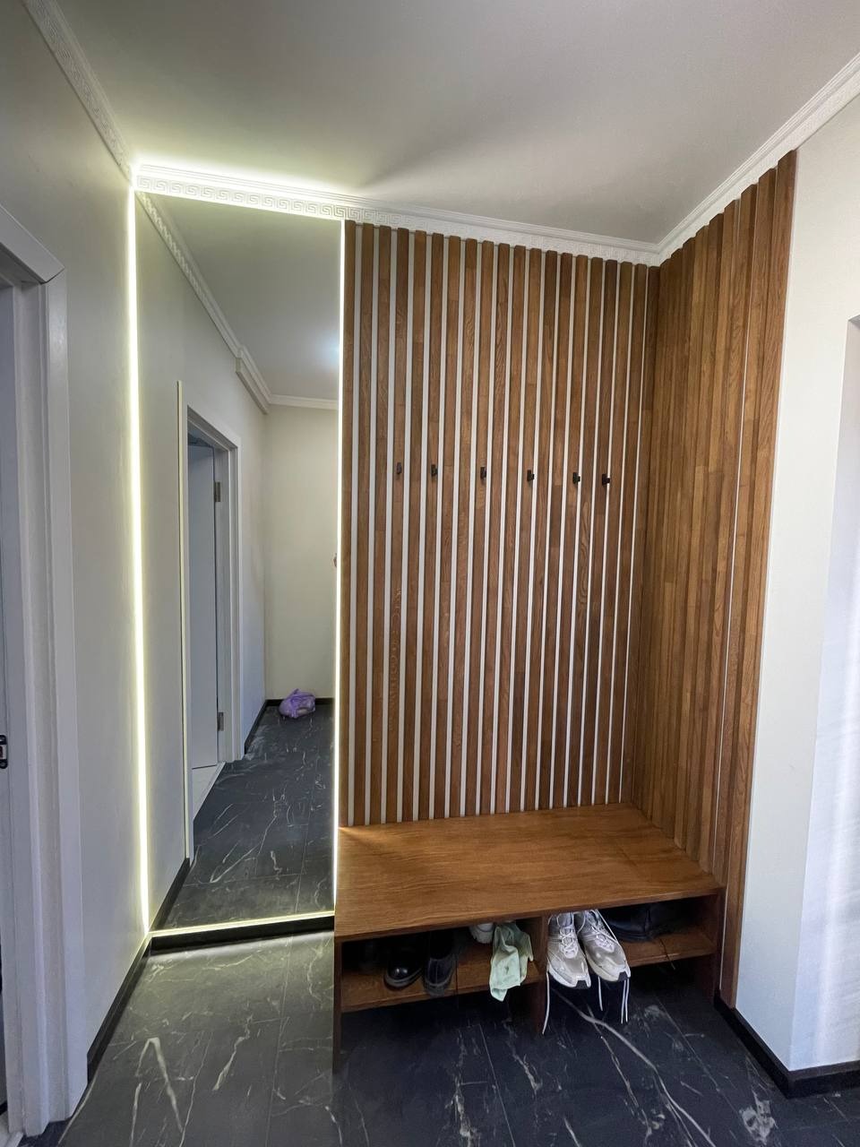 Nowoczesna aranżacja przedpokoju: drewniana boazeria z siedziskiem, lustro z podświetleniem LED, marmurowa podłoga. Minimalistyczny design, ciepłe oświetlenie.