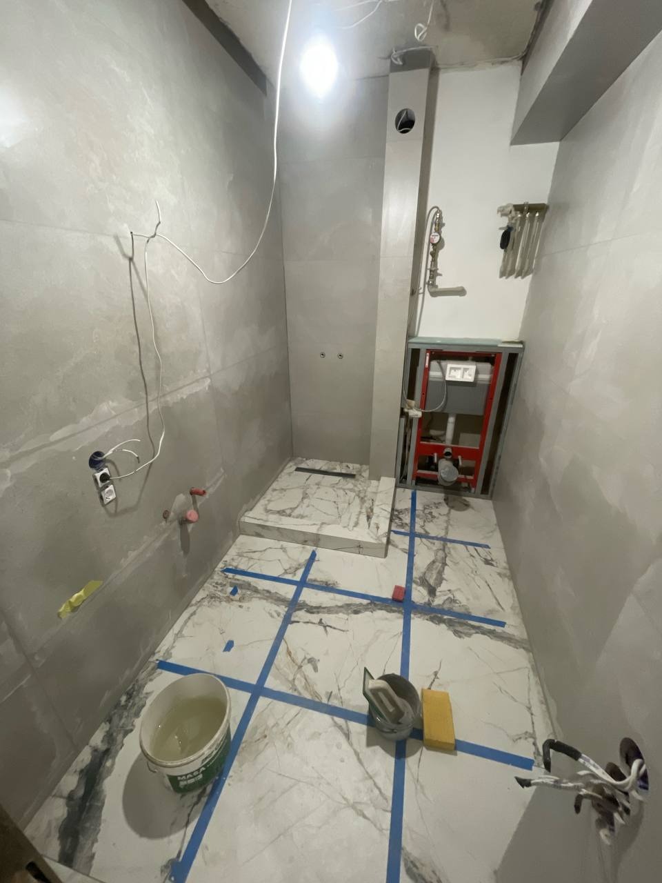 Wnętrze łazienki w trakcie prac wykończeniowych: ułożona marmurowa podłoga z niebieską taśmą, częściowo wykończone ściany, widoczne narzędzia i instalacje.