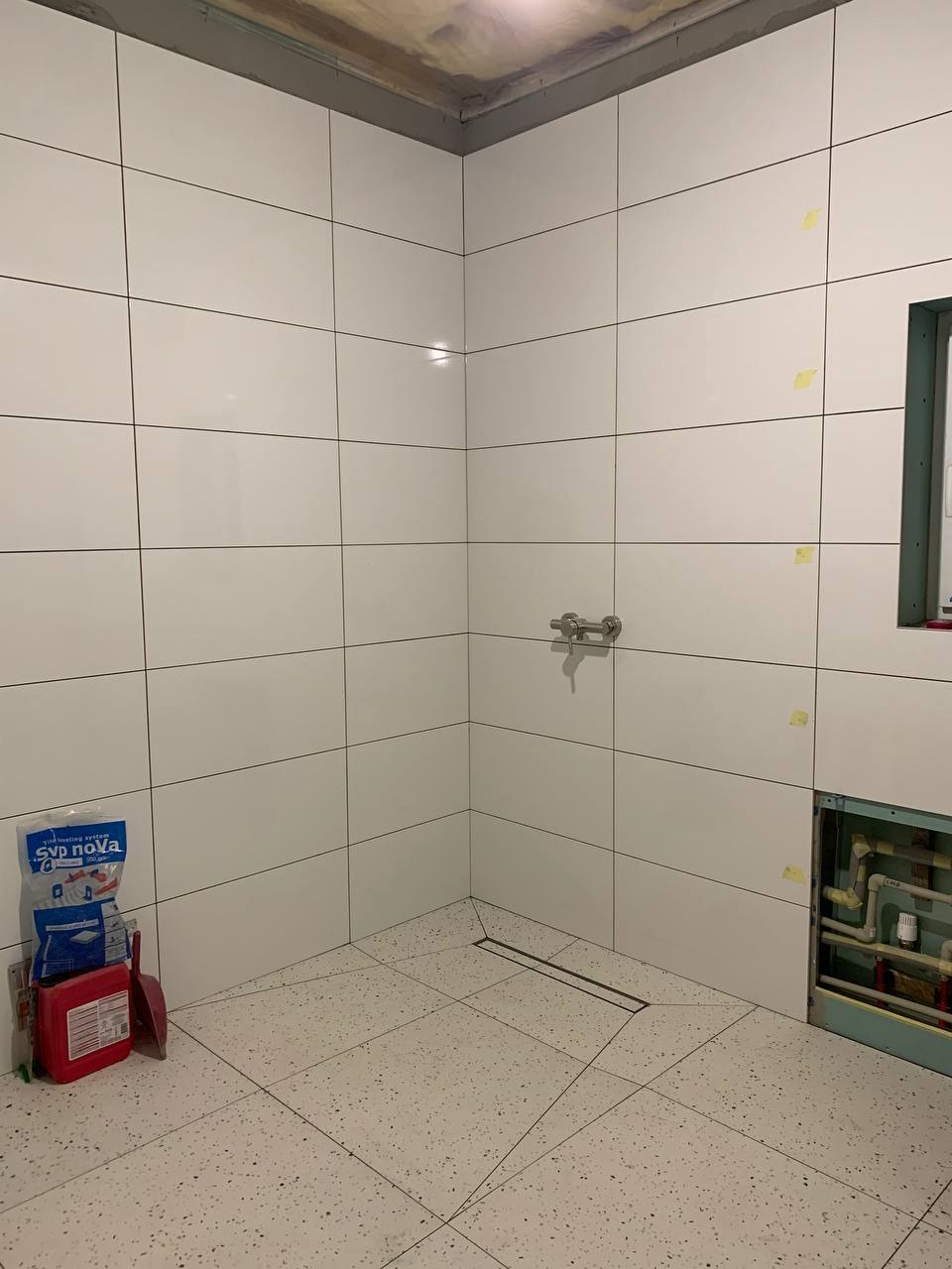 Wykończona łazienka z białymi, błyszczącymi płytkami na ścianach i podłodze w stylu lastryko. Widoczny odpływ liniowy i przygotowane miejsce na instalację.