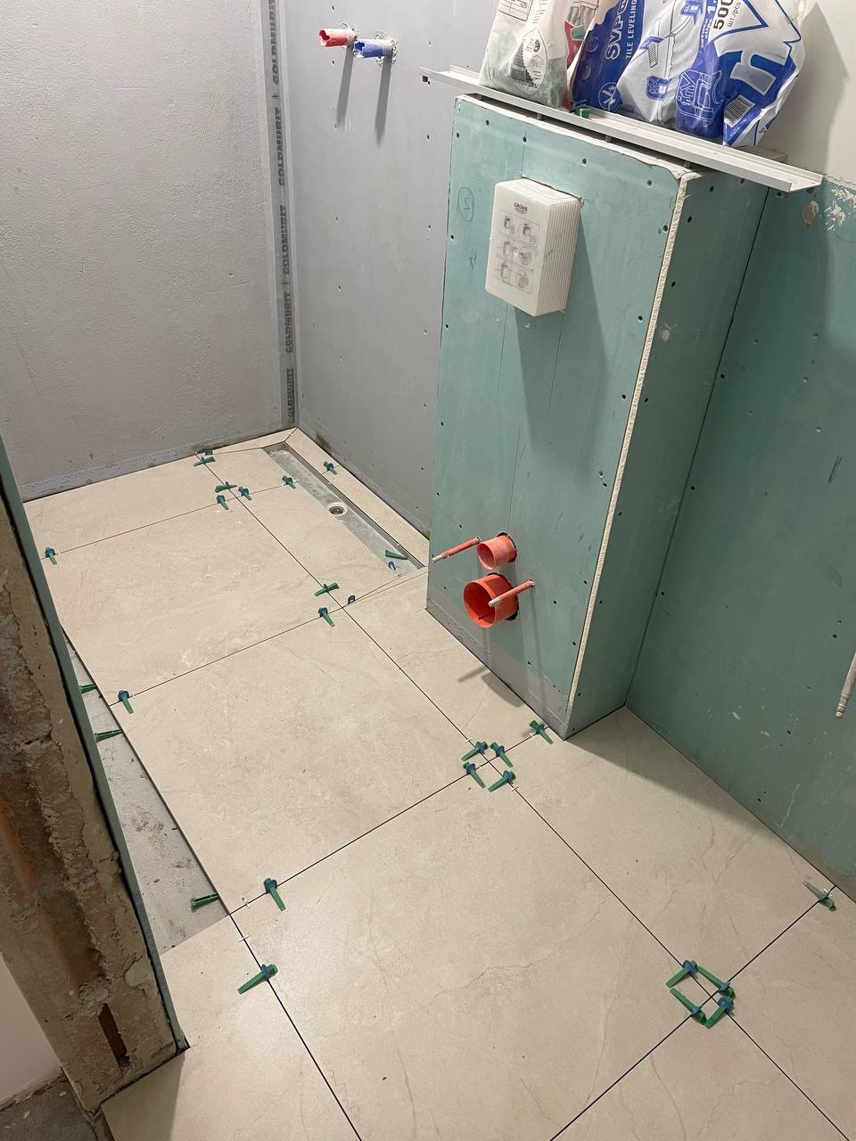 Układanie beżowych płytek podłogowych w łazience z użyciem systemu poziomowania. Widoczne elementy instalacji i płyta karton-gips na ścianie.