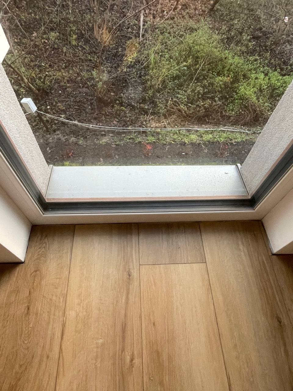 Widok z balkonu na zewnątrz, z drewnianą podłogą i fragmentem ściany z oknem. Na zewnątrz widoczna zieleń i krzewy.