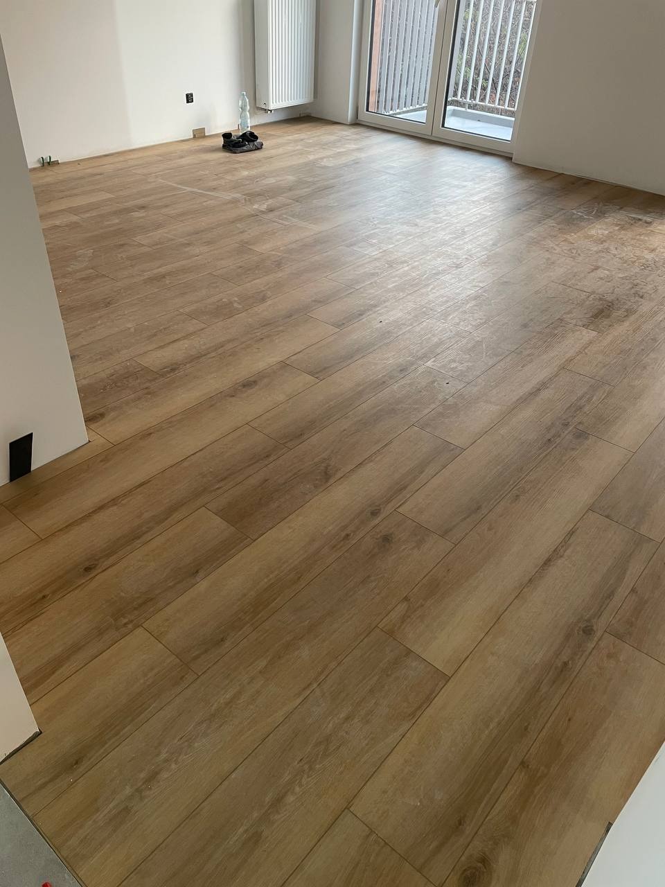 Ukończona podłoga z paneli imitujących drewno w jasnym, przestronnym pokoju z białymi ścianami i dużym oknem balkonowym. Widoczne detale wykończenia.