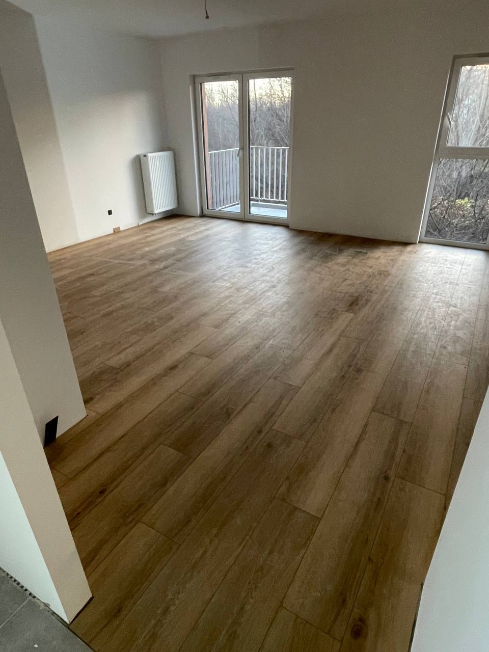 Jasne wnętrze z drewnopodobną podłogą, grzejnikiem i balkonem z widokiem na drzewa. Świeżo wykończone, minimalistyczne pomieszczenie z naturalnym światłem.