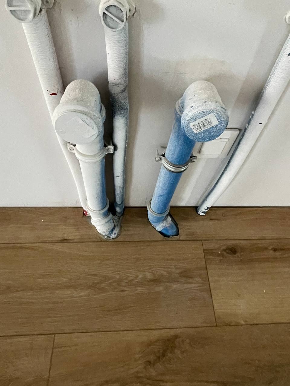 Pionowe rury, częściowo pomalowane na biało i niebiesko, przymocowane do ściany nad drewnianą podłogą. Widoczne ślady farby na podłodze i rurach.