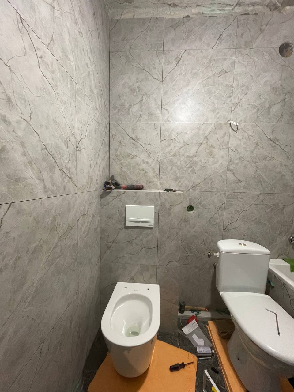 Łazienka w trakcie remontu: szare płytki imitujące marmur na ścianach, częściowo zamontowana toaleta podwieszana i stojąca, widoczne narzędzia i materiały budowlane.