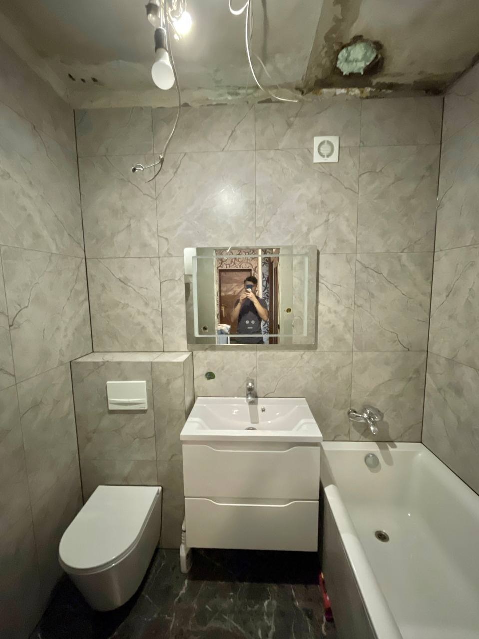 Wyremontowana łazienka z szarymi płytkami imitującymi marmur, białą armaturą i lustrem. Widoczne niedokończone prace na suficie i odbicie osoby robiącej zdjęcie.