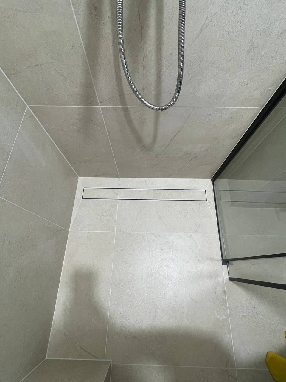 Wykończona kabina prysznicowa z odpływem liniowym, wyłożona jasnymi płytkami imitującymi marmur. Widoczny fragment szklanej ścianki i węża prysznicowego.