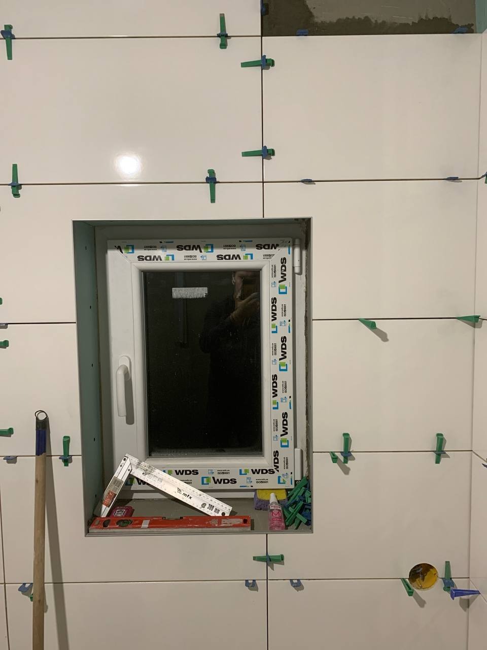 Białe płytki ścienne w trakcie układania wokół okna, z użyciem poziomicy i krzyżyków dystansowych. Widoczne narzędzia i materiały budowlane.