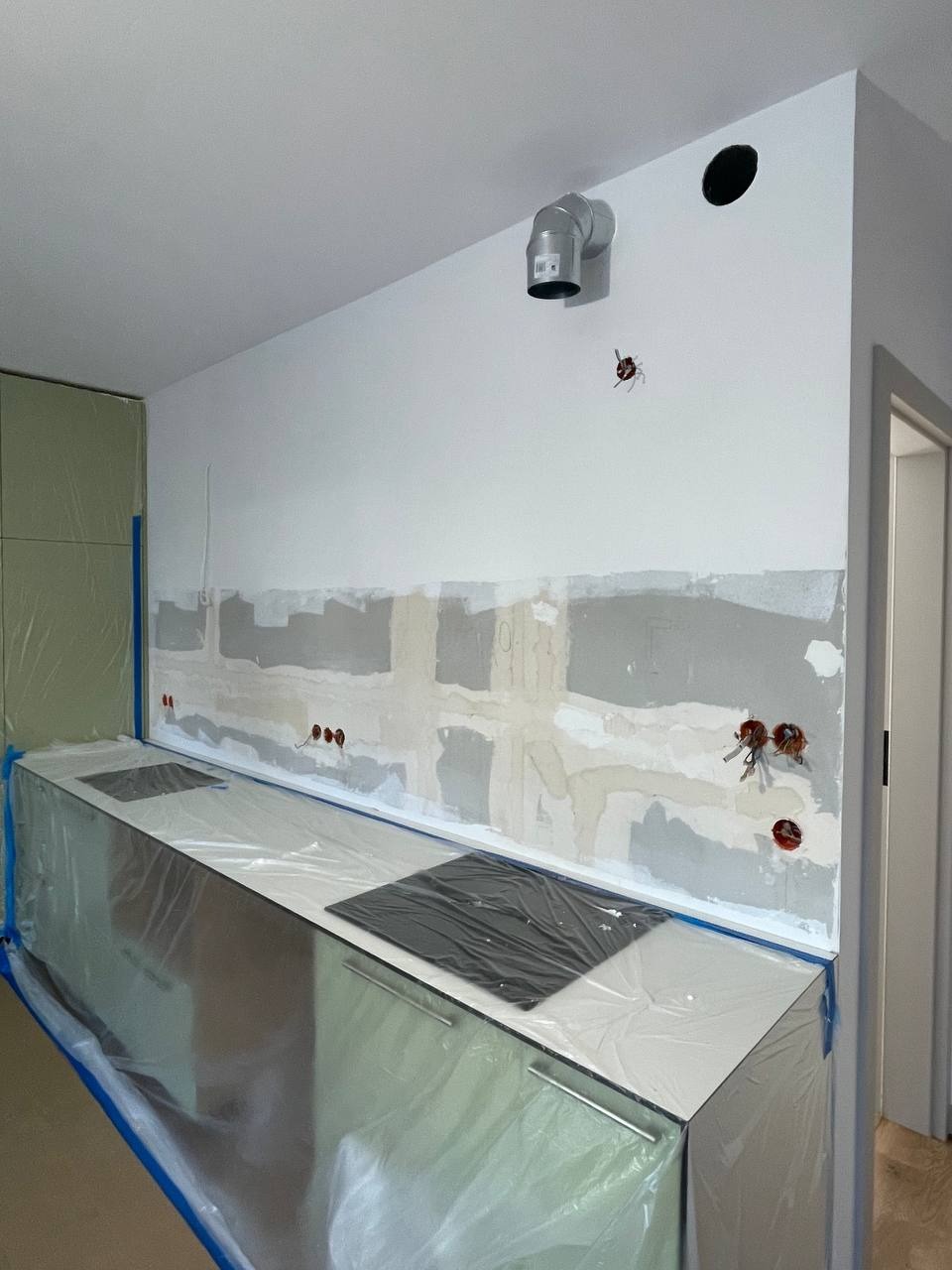 Kuchnia w trakcie remontu: ściana po szpachlowaniu, zabezpieczone folią meble kuchenne, widoczne instalacje elektryczne i wentylacyjne.