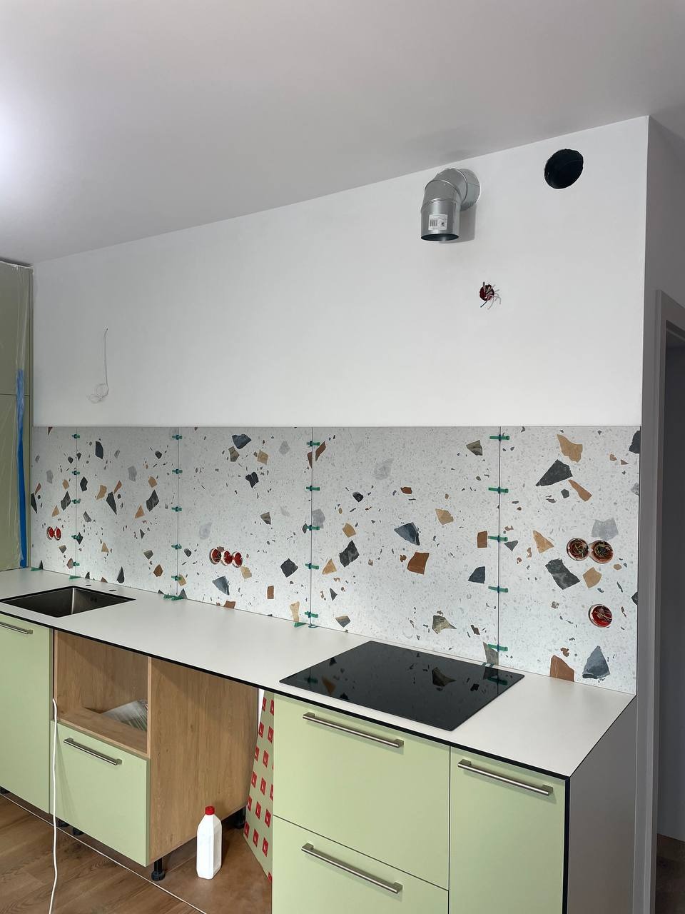 Kuchnia w trakcie wykończenia: zielone szafki, biały blat, ściana z płytkami w stylu lastryko z krzyżykami dystansowymi. Widoczne gniazdka i otwór wentylacyjny.