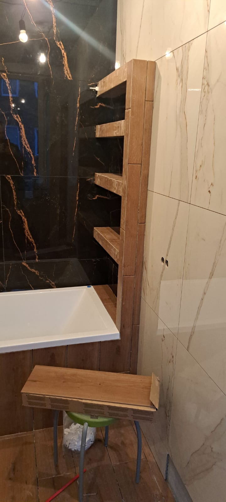 Wnętrze łazienki w trakcie remontu: czarne i beżowe płytki imitujące marmur, biała wanna, drewniane półki w zabudowie, taboret z narzędziami. Widoczne detale wykończenia.