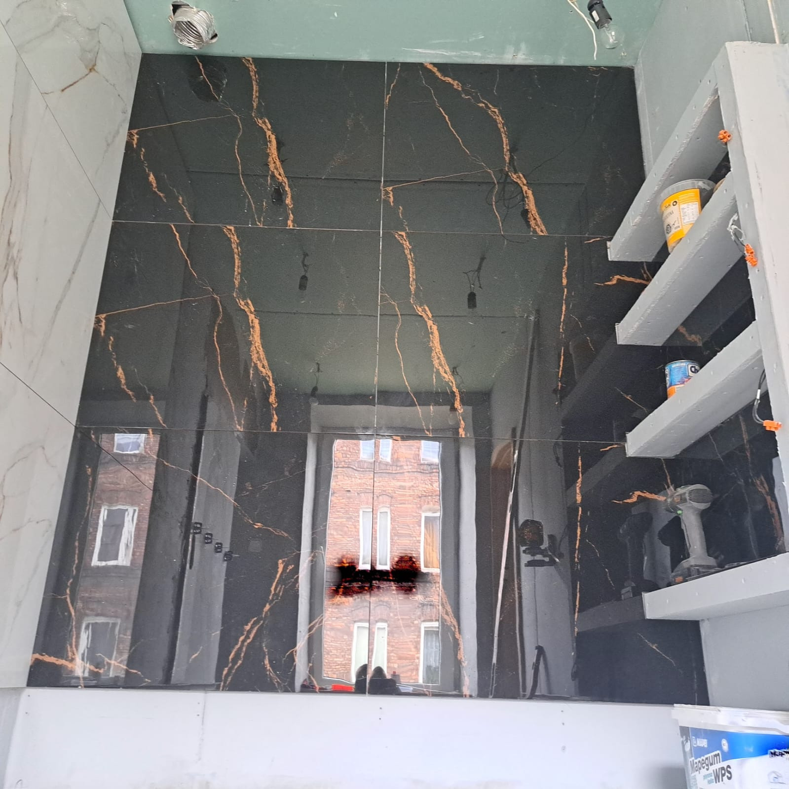 Ściana w łazience wyłożona czarnymi, błyszczącymi płytkami z marmurkowym, złotym wzorem. Widoczne półki i narzędzia, co sugeruje etap prac wykończeniowych.