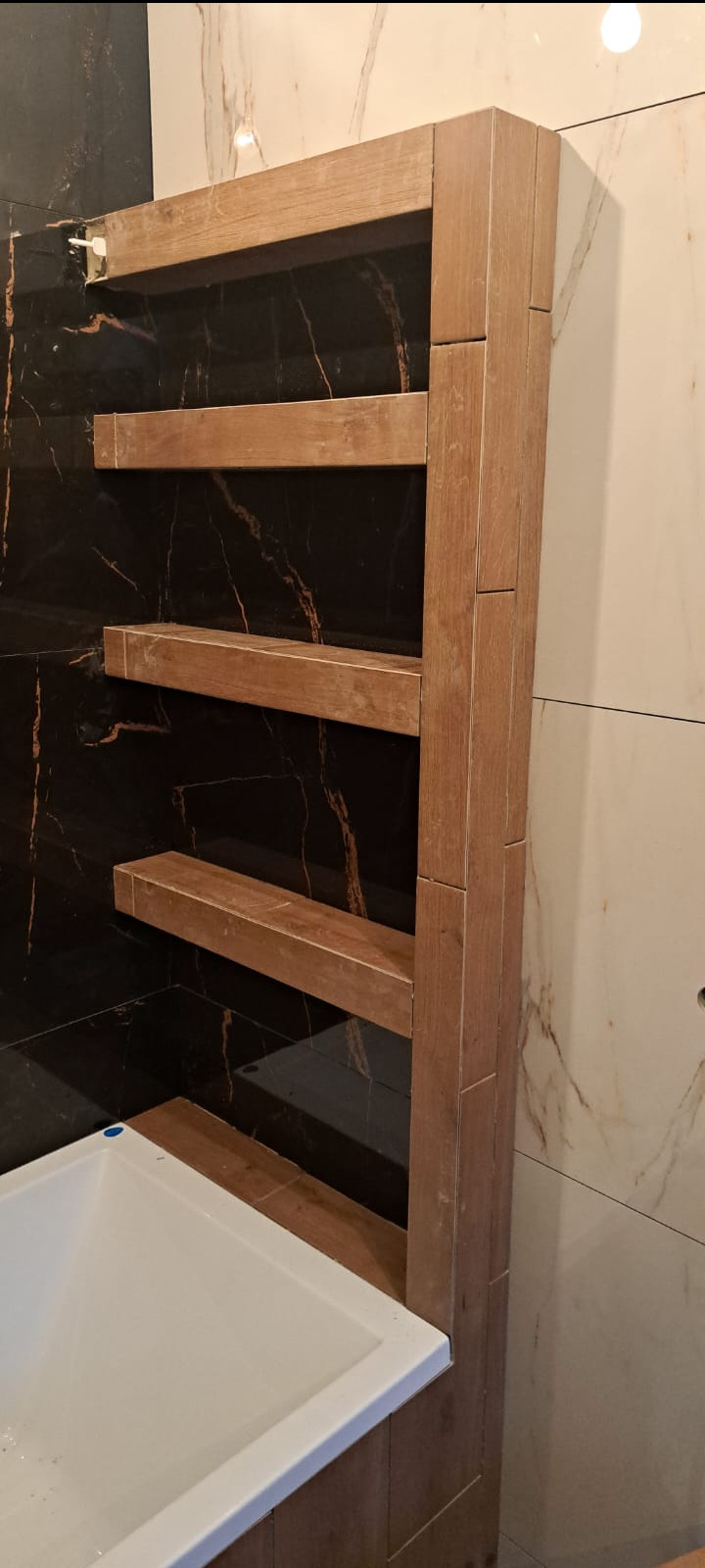 Wykończona łazienka z drewnianym regałem na ręczniki obok wanny, na tle marmurowych płytek w odcieniach czerni i beżu. Widoczne detale wykończenia.