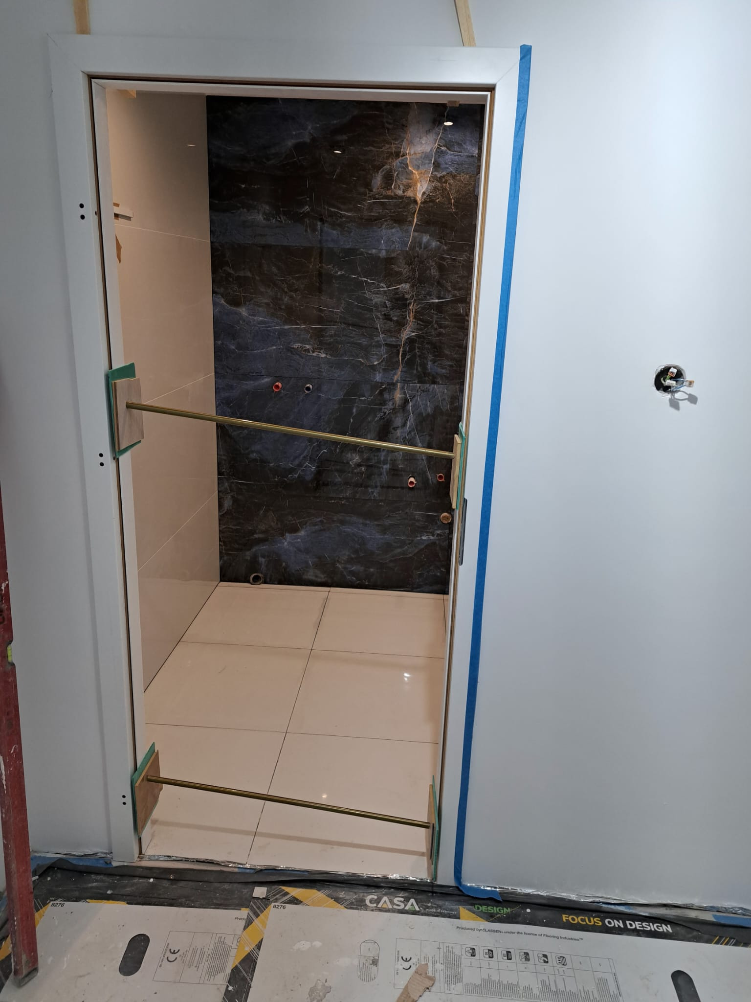 Wnętrze łazienki w trakcie wykończenia: widoczne płytki imitujące marmur na ścianie, białe płytki podłogowe i zabezpieczone drzwi z ościeżnicą.