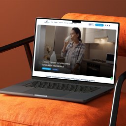 IdeaNOW - Tomasz Świercz - Laptop na pomarańczowym fotelu prezentuje stronę internetową Trade Milk z kobietą pijącą mleko w tle kuchni. Minimalistyczny design, czcionki czytelne, nawigacja intuicyjna.