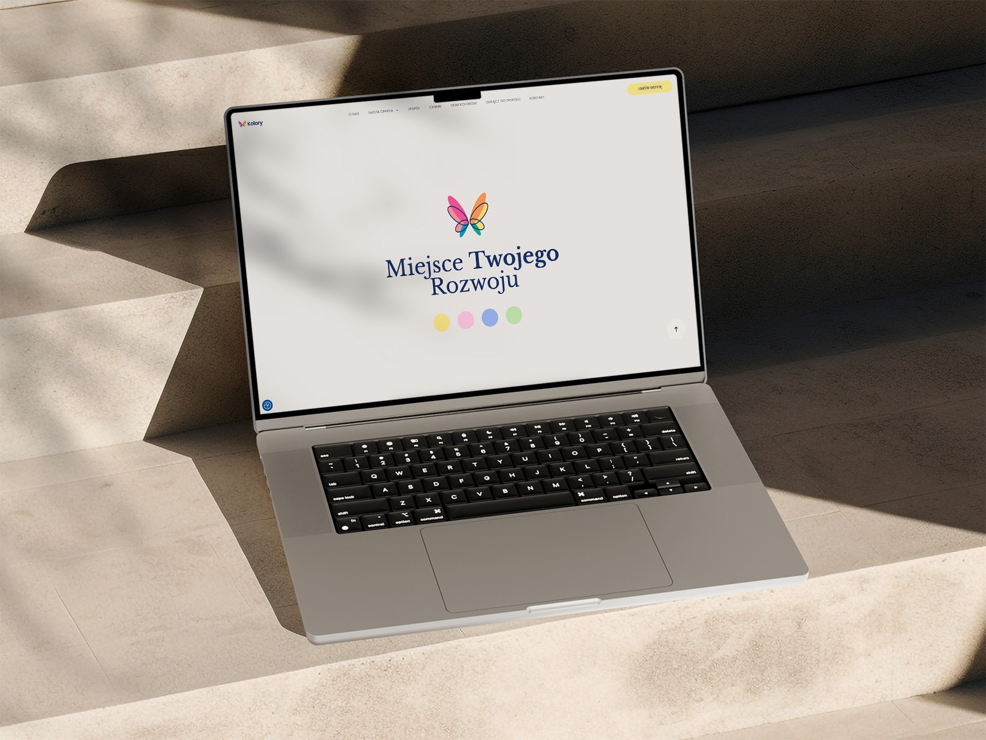 Laptop na schodach prezentuje stronę z logo motyla i napisem 'Miejsce Twojego Rozwoju', w stonowanej kolorystyce, oświetlony naturalnym światłem.