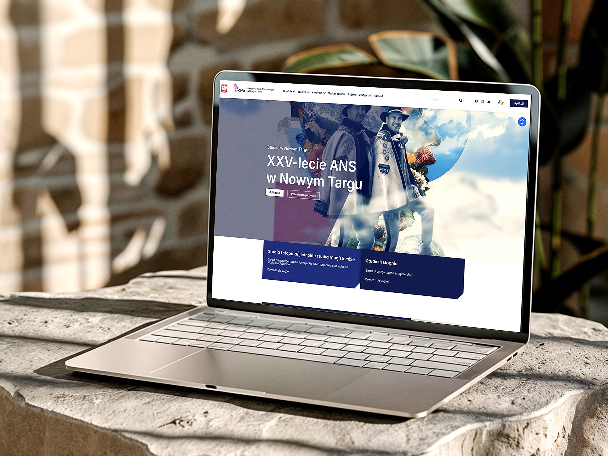 Laptop na kamiennym blacie prezentuje stronę internetową Akademii Nauk Stosowanych w Nowym Targu, z motywem XXV-lecia uczelni i górali.