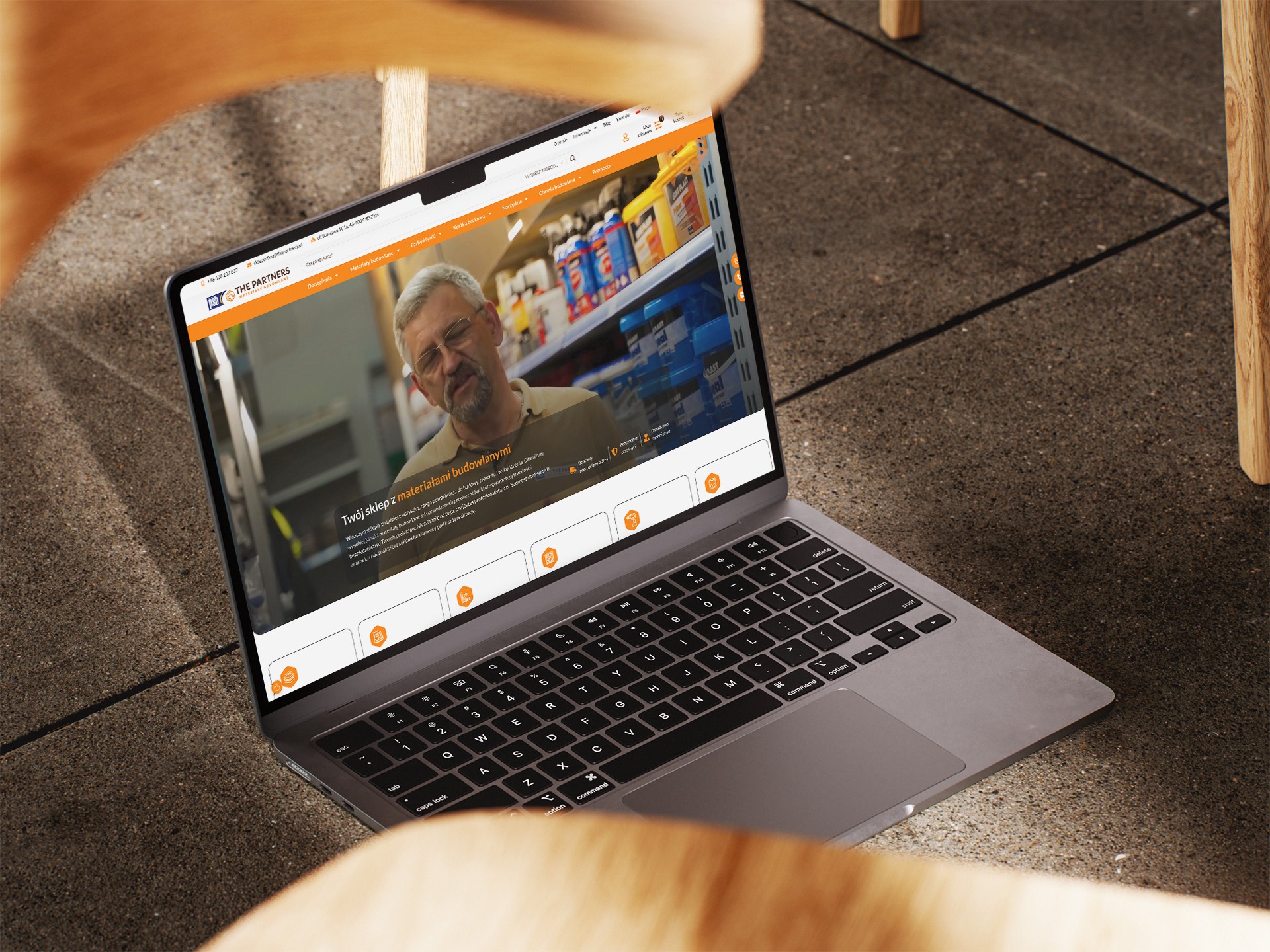 Laptop wyświetla stronę internetową sklepu budowlanego 'The Partners', z widocznym nagłówkiem, menu i fragmentem oferty. Ujęcie z perspektywy siedzącego, przez oparcie krzesła.