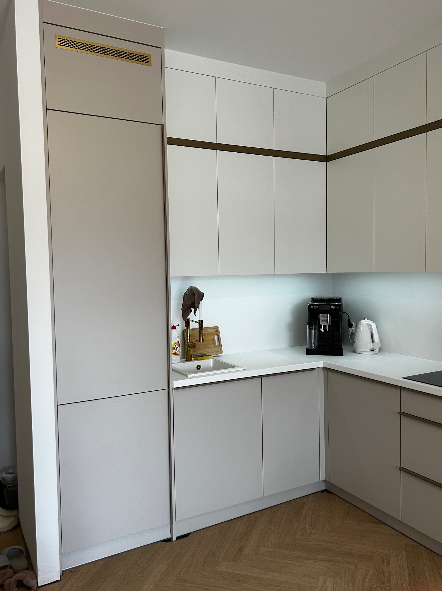 Jasnoszara kuchnia z zabudową pod sufit, złote akcenty na szafkach, drewniana podłoga w jodełkę. Na blacie ekspres do kawy i czajnik.