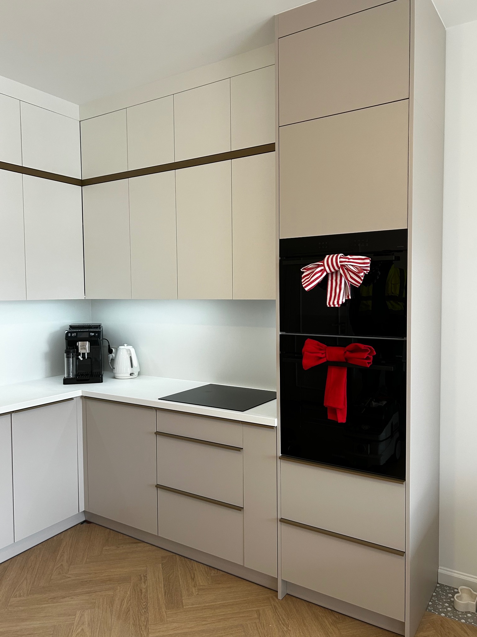 Jasna kuchnia na wymiar z beżowymi frontami i złotymi uchwytami. Na piekarniku świąteczne kokardy. Ekspres do kawy i czajnik na blacie.