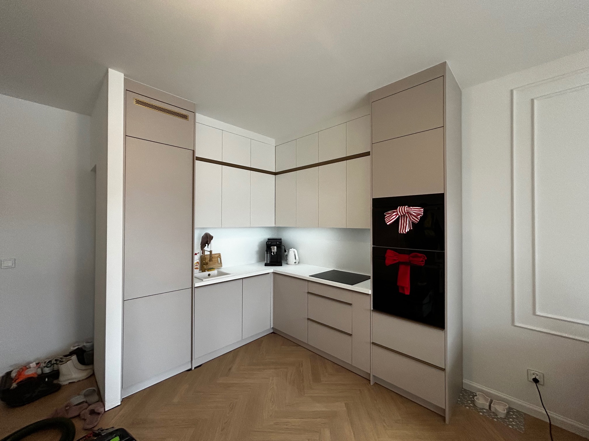 Jasna kuchnia w zabudowie z beżowymi akcentami i drewnianą podłogą w jodełkę. Na piekarniku dekoracja w postaci czerwonych kokardek.