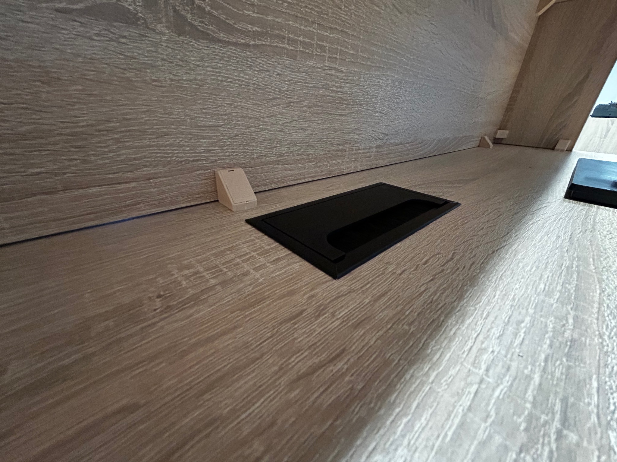 Fragment wnętrza szafy z jasnego drewna z wbudowanym czarnym gniazdem elektrycznym i zaślepką, ukazujący detale wykończenia i funkcjonalne rozwiązania.