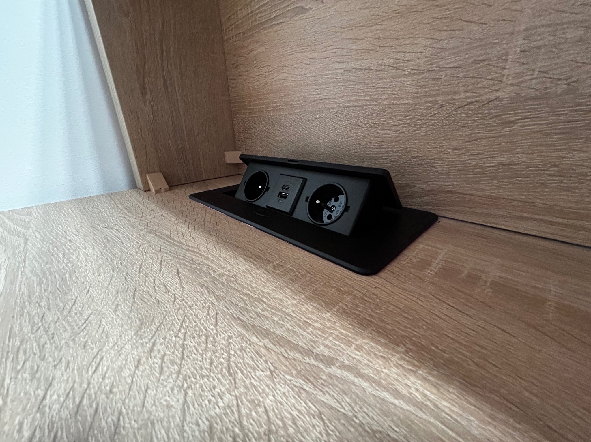 Wpuszczane gniazdko elektryczne z portami USB, zamontowane w blacie meblowym o drewnopodobnym wykończeniu. Widoczne detale montażu wewnątrz szafki.