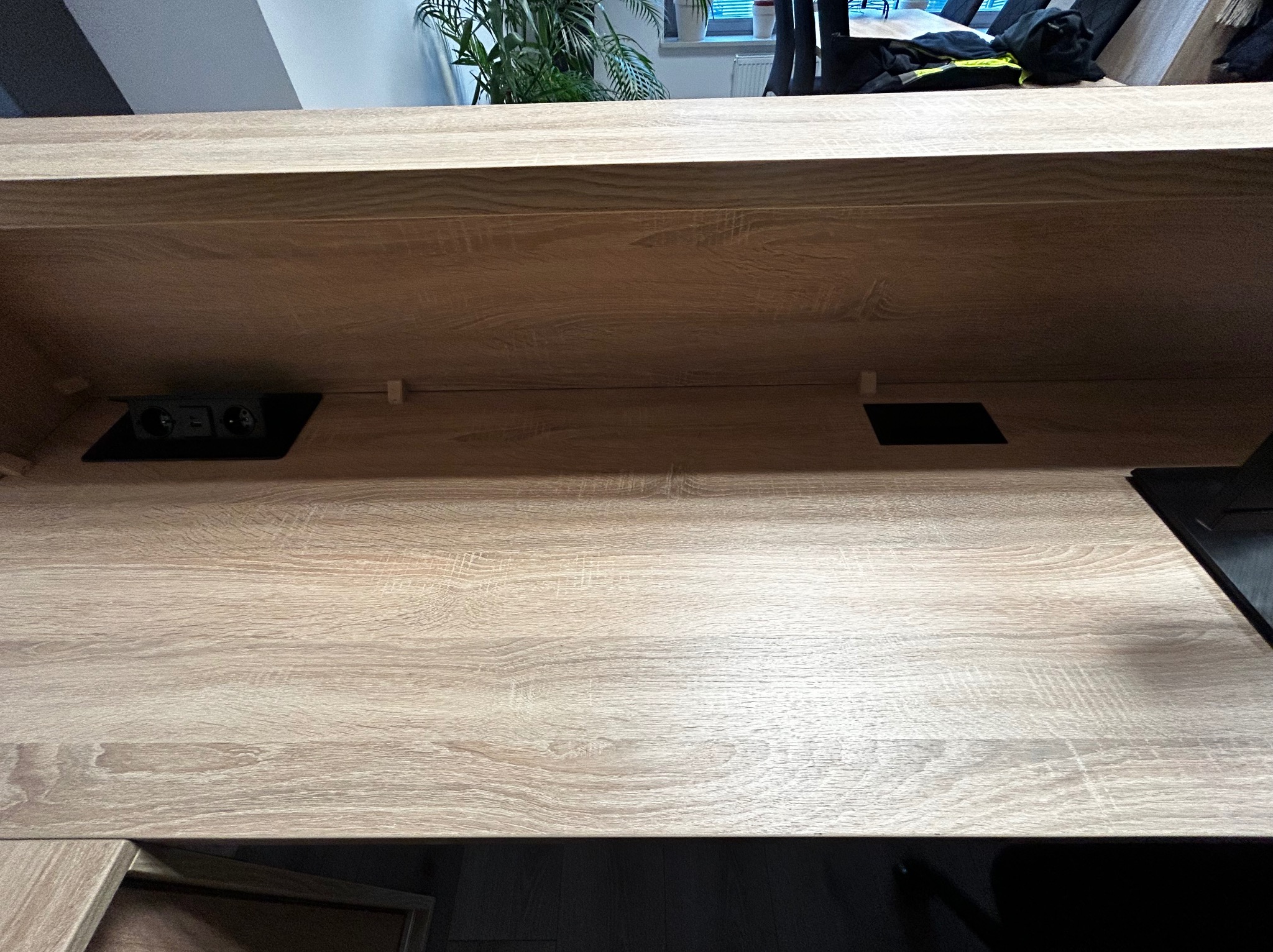 Blat roboczy z jasnego drewna z gniazdkami elektrycznymi i otworem, prawdopodobnie element kuchni na wymiar. Widok z dołu, fragment rośliny w tle.