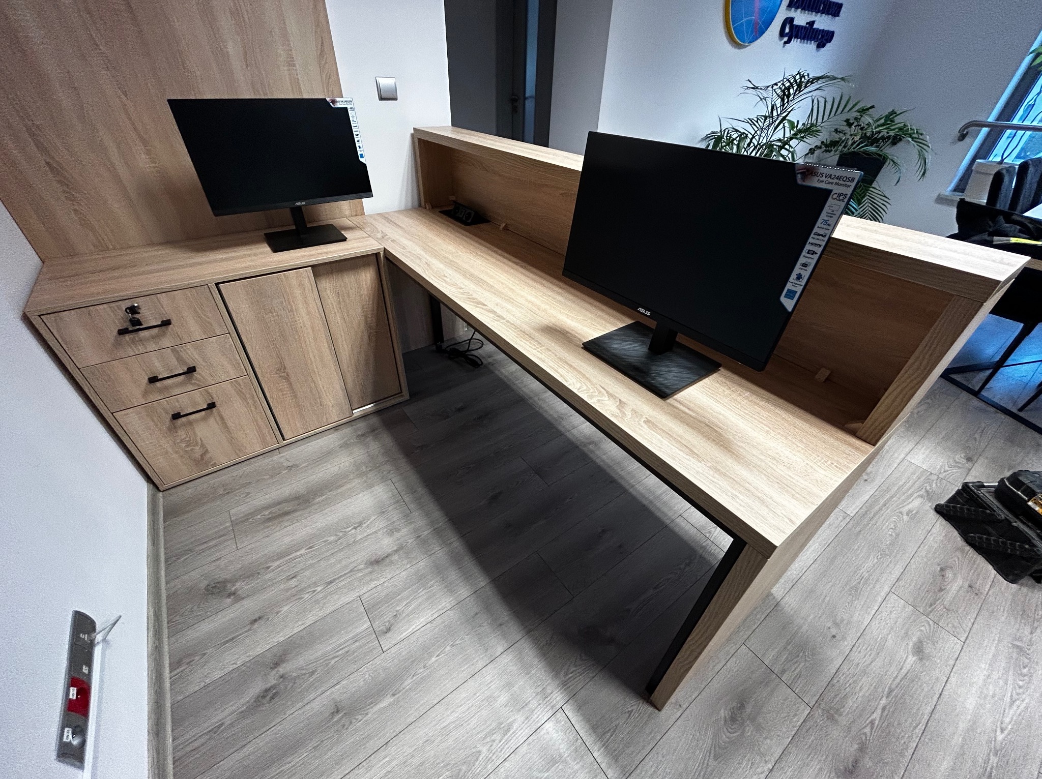 Jasna, drewniana lada recepcyjna z szafkami i dwoma monitorami. Nowoczesny design, widoczne detale wykończenia i czarne nogi. Ustawiona w jasnym pomieszczeniu biurowym.